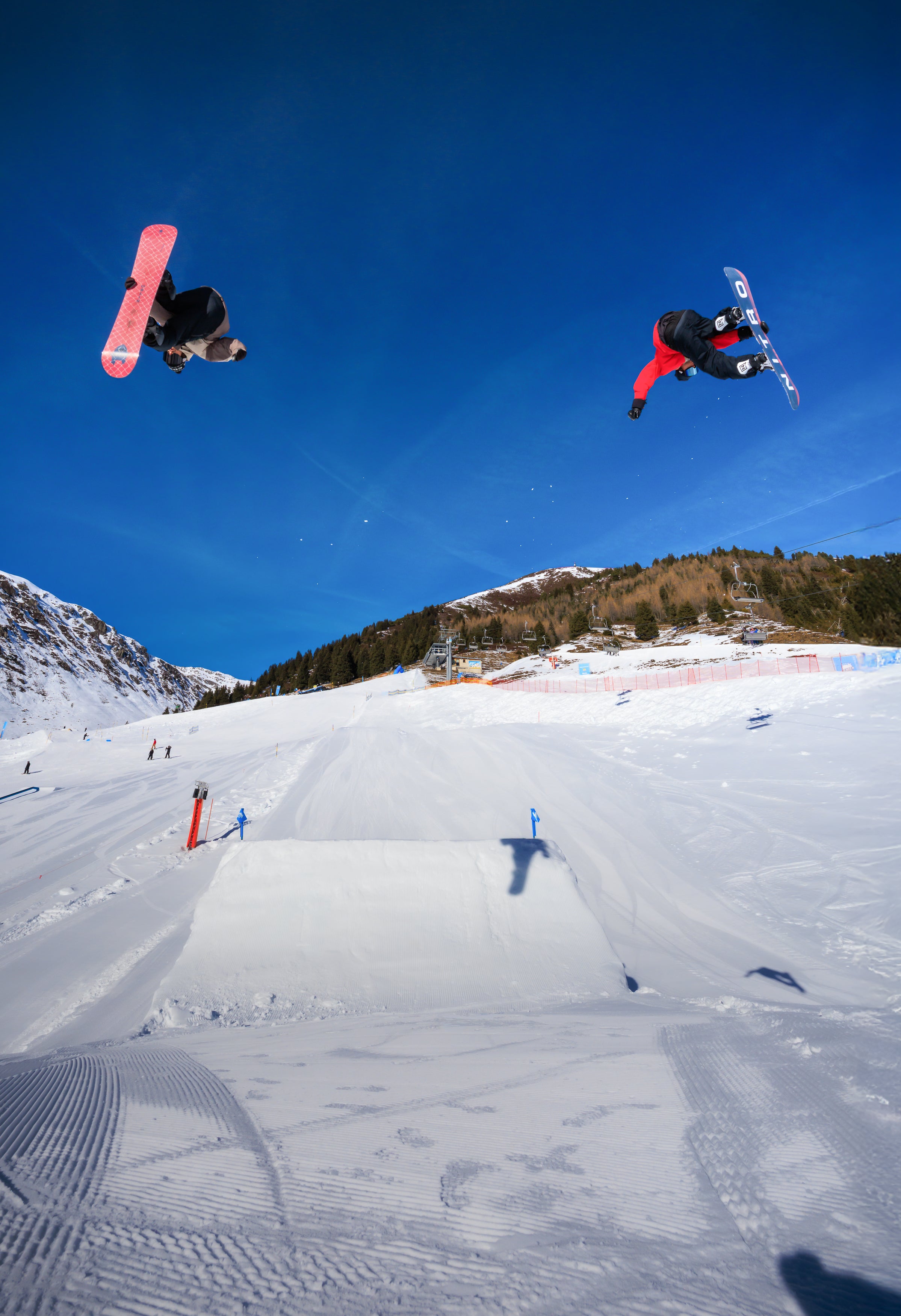 Two snowboarders performing aerial tricks above a snowy slope with mountains in the background.