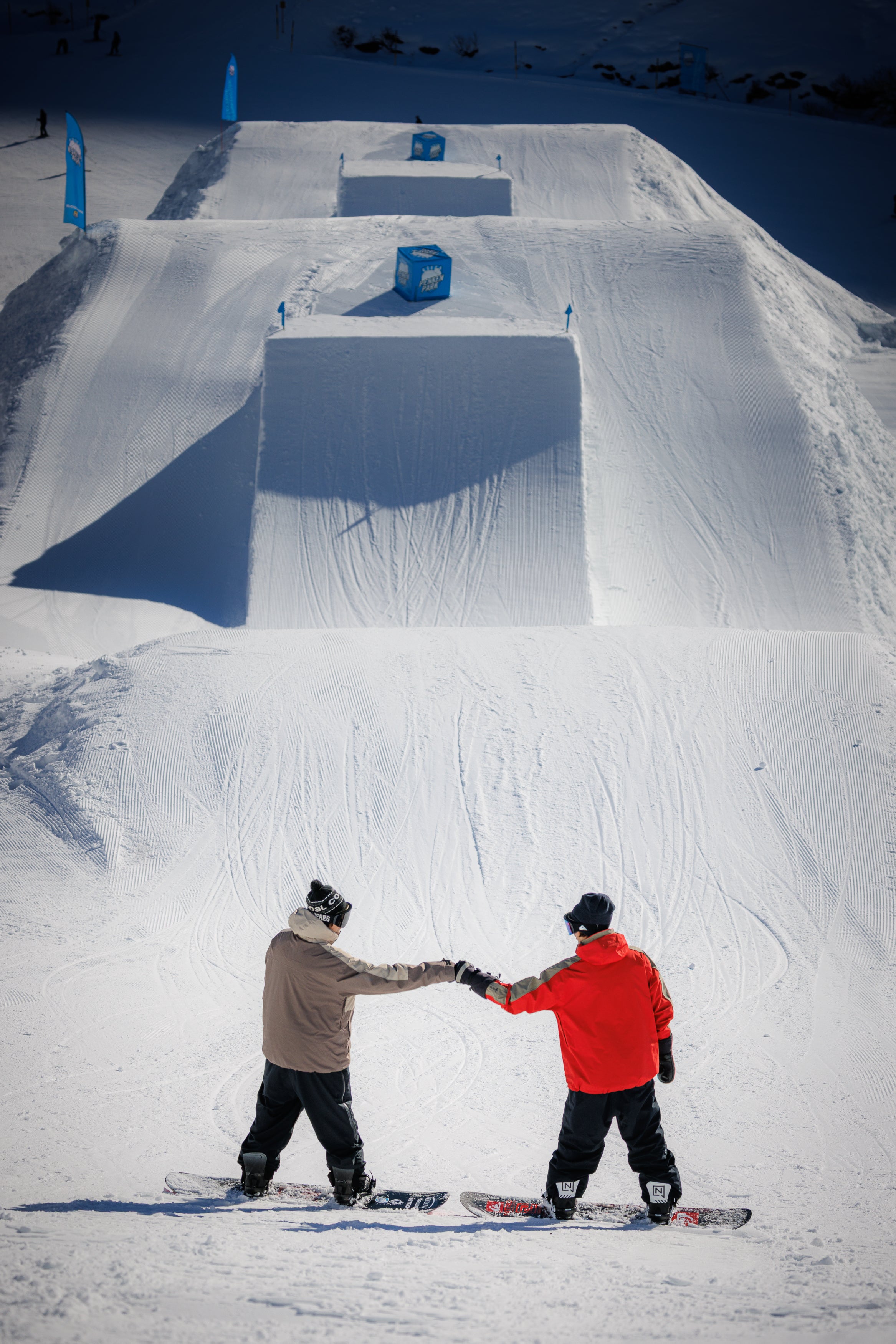 Tom and Werni fist bumping on a snowy slope with jumps and obstacles.